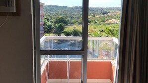 Una habitación con un gran ventanal corredera que da a un balcón. El balcón tiene barandilla metálica y vistas a un paisaje verde con árboles y un edificio distante.