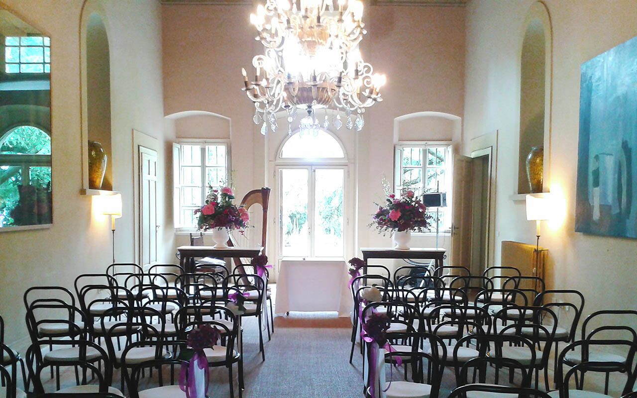 A room with rows of black chairs facing a central aisle, a chandelier, and two vases with flowers on tables.
