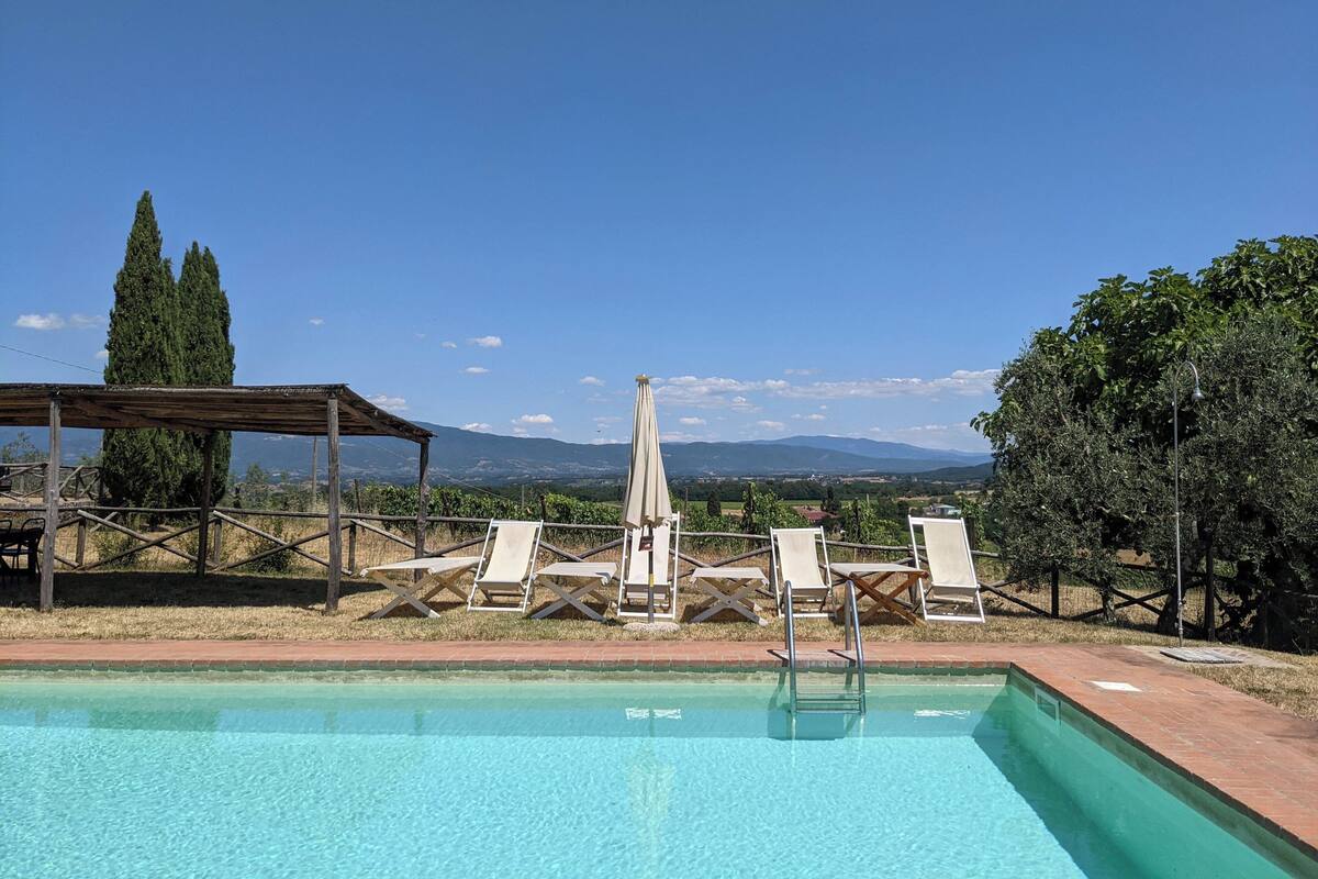 A pool area with lounge chairs and a pergola, surrounded by mountains and greenery.