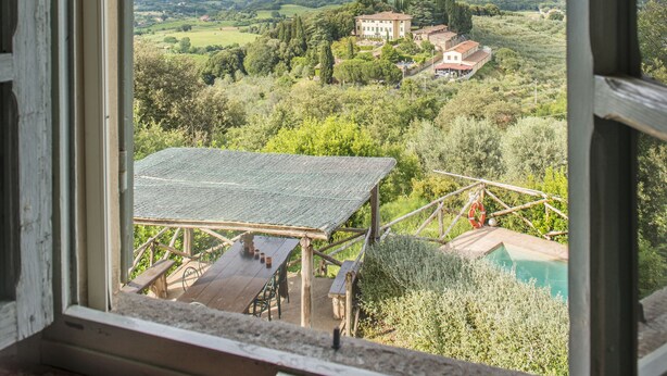 A view through a window of a countryside landscape with a wooden deck, a swimming pool, and distant buildings. - Petrolo (Bucine)
