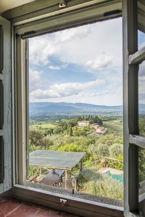 A view through a window of a countryside landscape with a wooden deck, a swimming pool, and distant buildings. - Petrolo (Bucine)