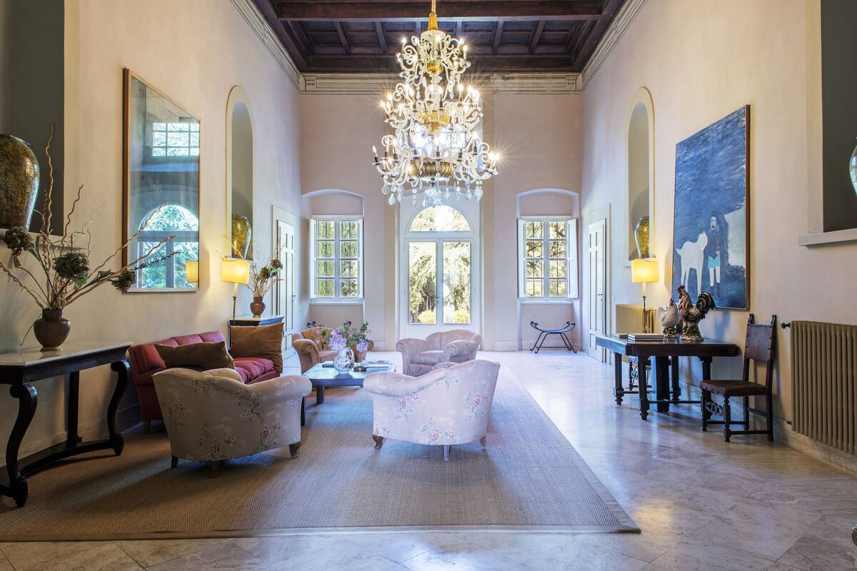 A spacious living room with a chandelier, floral armchairs, a coffee table, and a large painting on the wall.