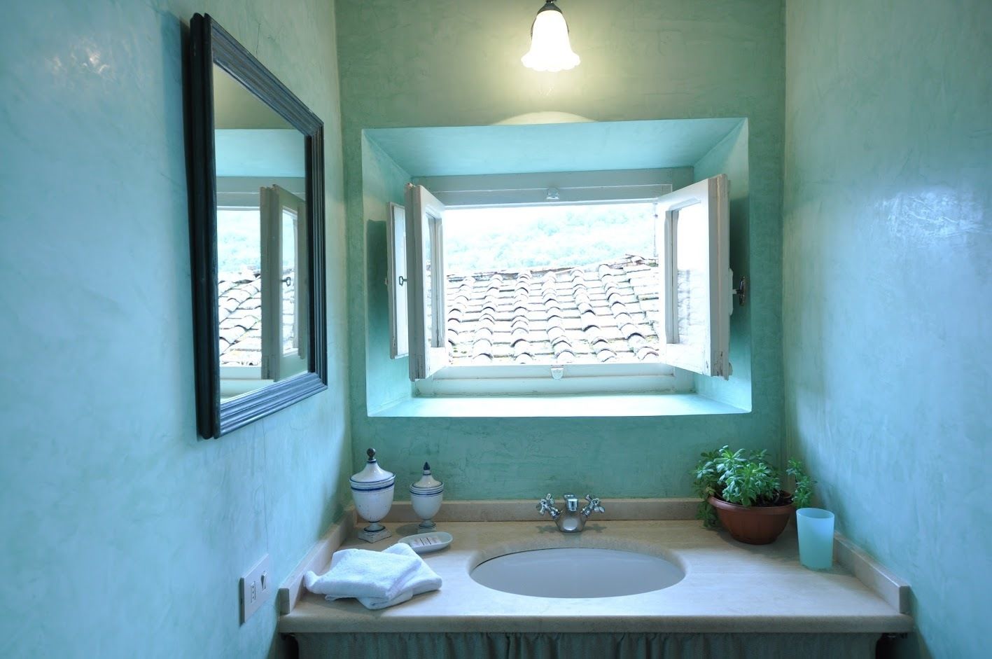 A bathroom with a sink, a window with open shutters, a mirror, and a light fixture.