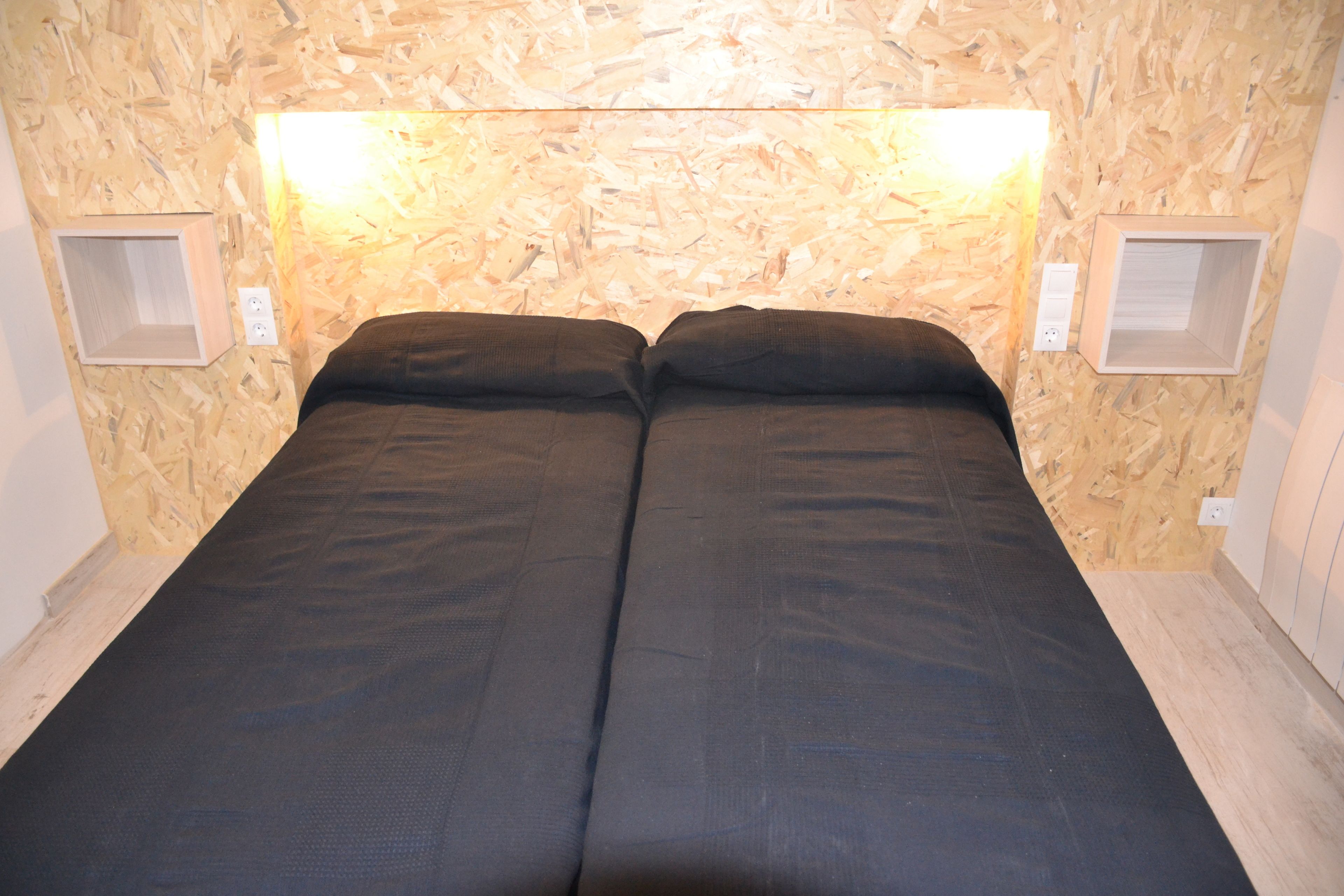 A room with a wooden wall, a bed with black bedding, and two wall-mounted light fixtures.