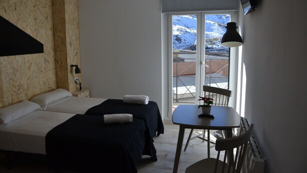 A room with two beds, a table with a chair, a lamp, and a view of snow-capped mountains through a glass door. - Gorbea Suites (Monachil)