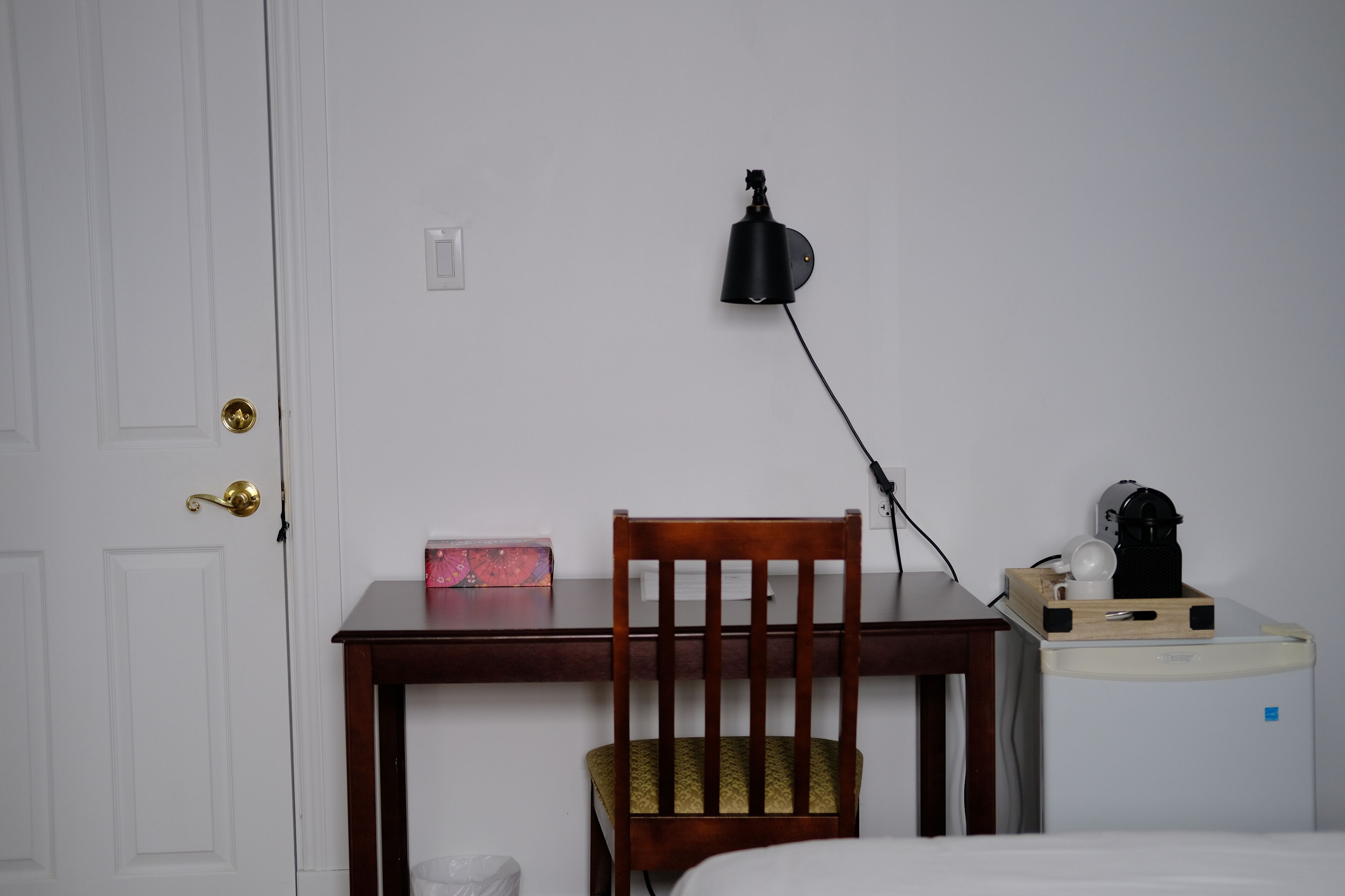 A room with a wooden desk, a chair, a lamp, a coffee machine, and a bed.