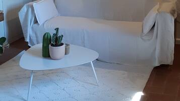 Ein gemütliches Wohnzimmer mit einem weißen Sofa, einem weißen Couchtisch und einem gemusterten Teppich. An den Wänden befinden sich Holzregale mit Pflanzen und Büchern.