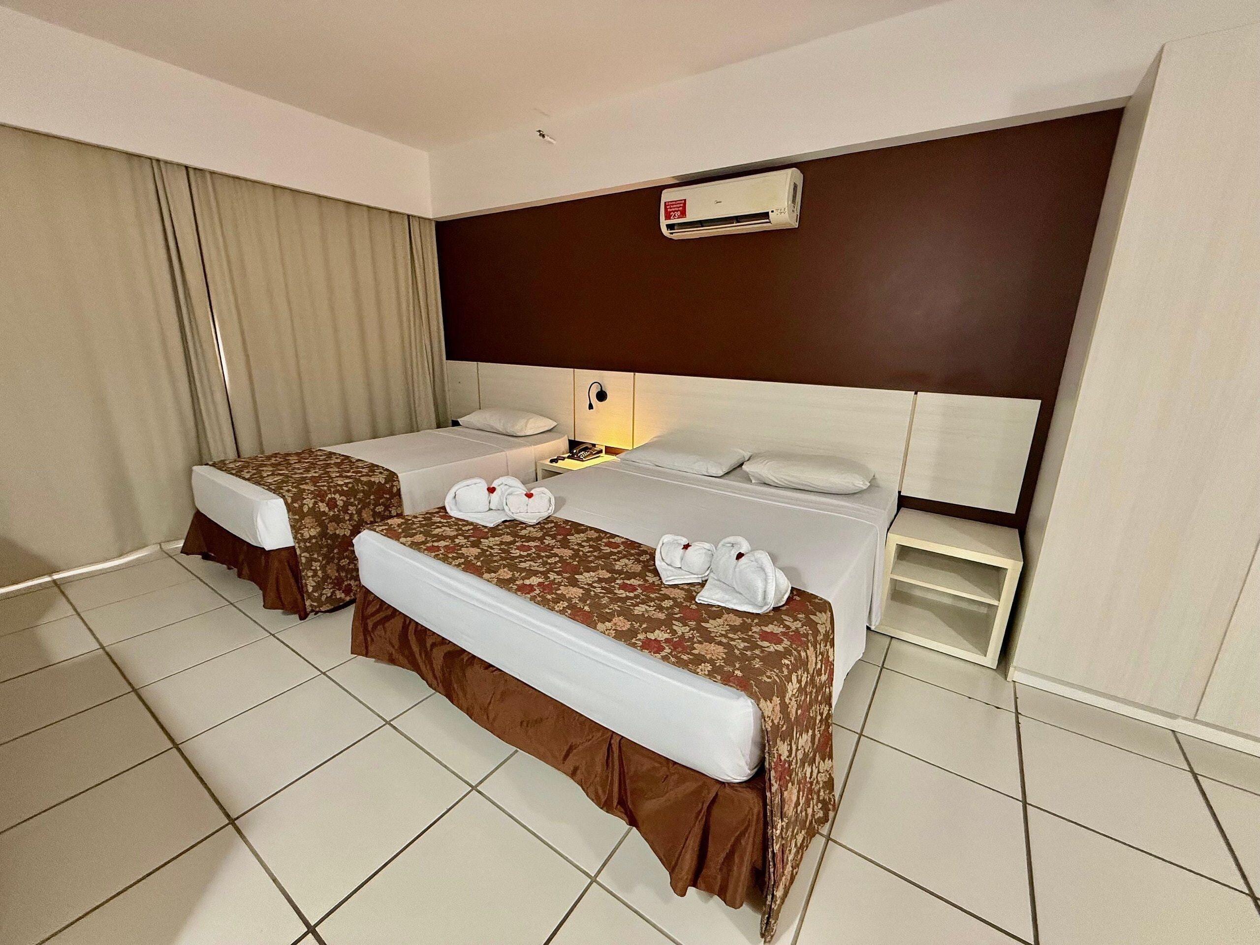 A hotel room with two beds, a nightstand, an air conditioner, and a window with curtains.
