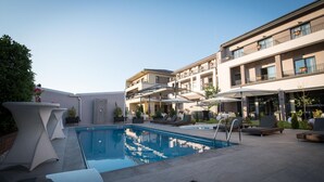 Zona de piscina con sillas de descanso y sombrillas, rodeada por un edificio con balcones y mesas al aire libre.