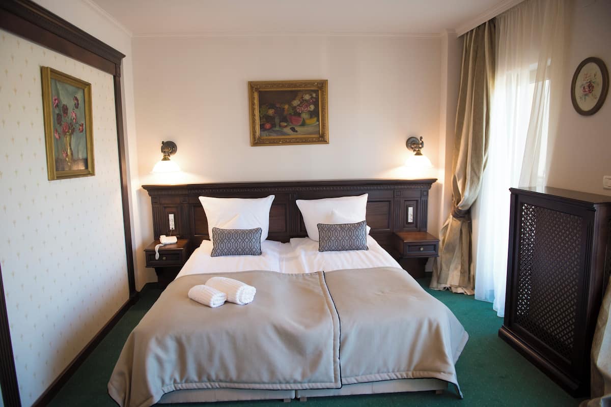 Un cuarto de hotel con una cama grande, dos mesitas de noche, una lámpara, un cuadro en la pared y una ventana con cortinas.