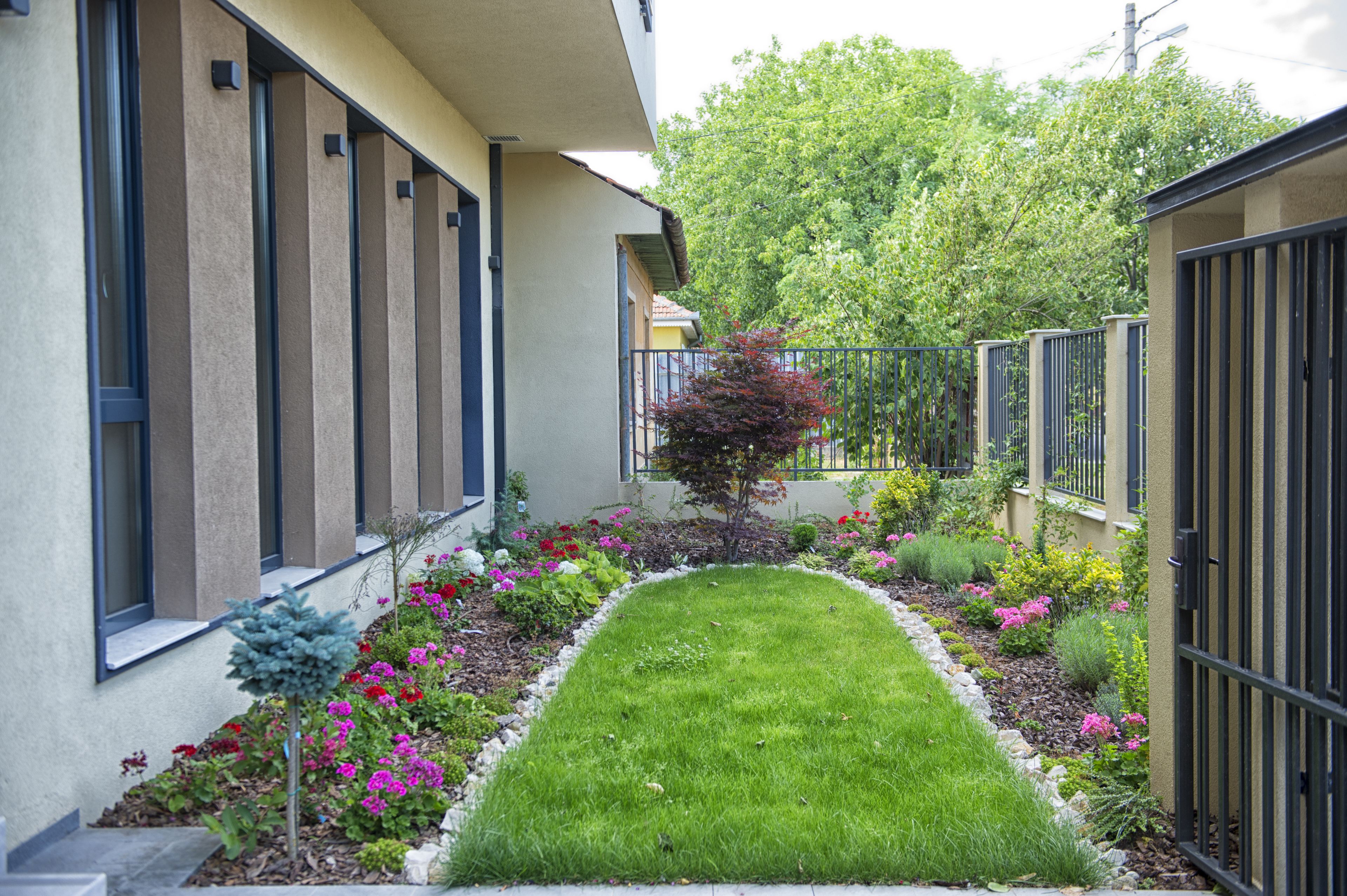 A residential building with a narrow, paved pathway leading to the entrance, surrounded by a well-maintained garden with various plants and flowers.