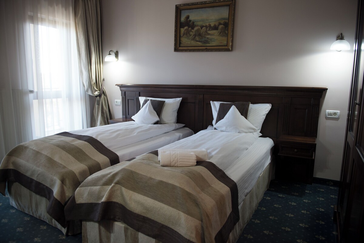 A hotel room with two beds, a painting on the wall, and a window with curtains.