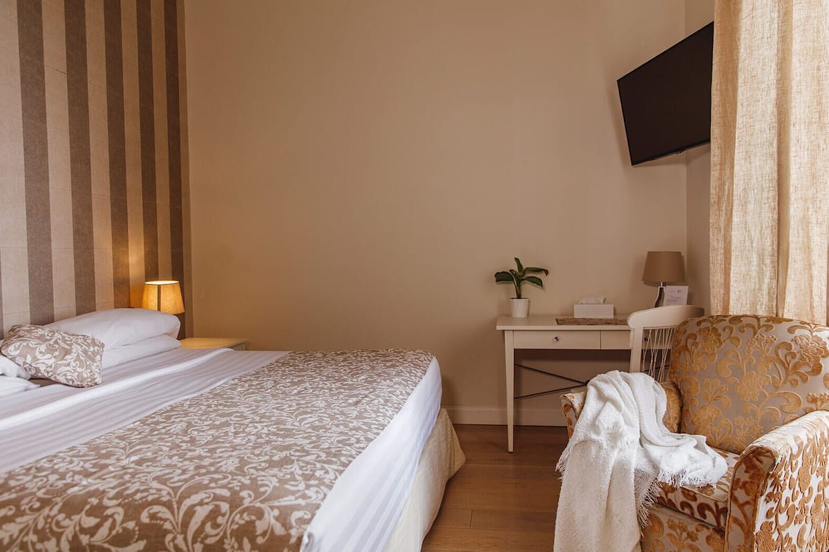 A hotel room with a bed, a desk, a chair, a TV mounted on the wall, and a potted plant.