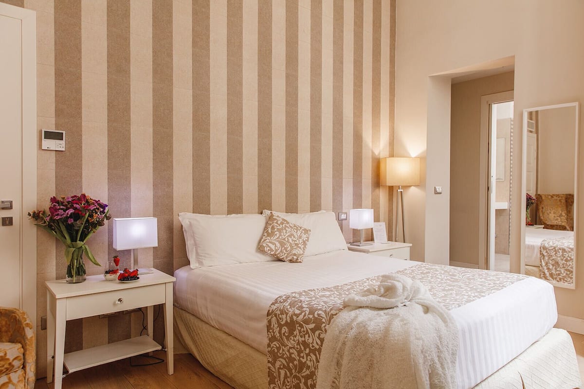 A hotel room with a striped wallpaper, a bed with white linens, two bedside tables with lamps, a vase of flowers, and a mirror.