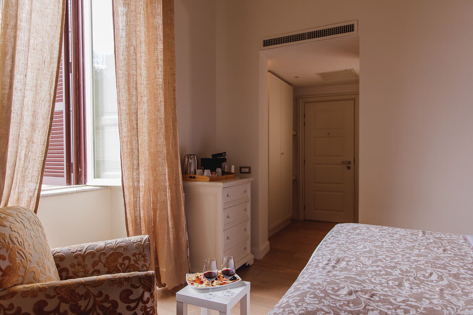 A bedroom with a bed, a dresser, a chair, and a small table with wine glasses and a plate of food.