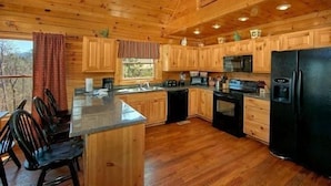 A wooden cabin kitchen with a central island, wooden cabinets, a black refrigerator, and a window with a view of trees. - Smoky Mountain Lodge (Pigeon Forge)