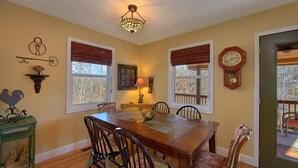 A dining room with a wooden table and chairs, two windows with blinds, a wall clock, and a decorative wall piece. - Gettin Cozy (Sevierville)