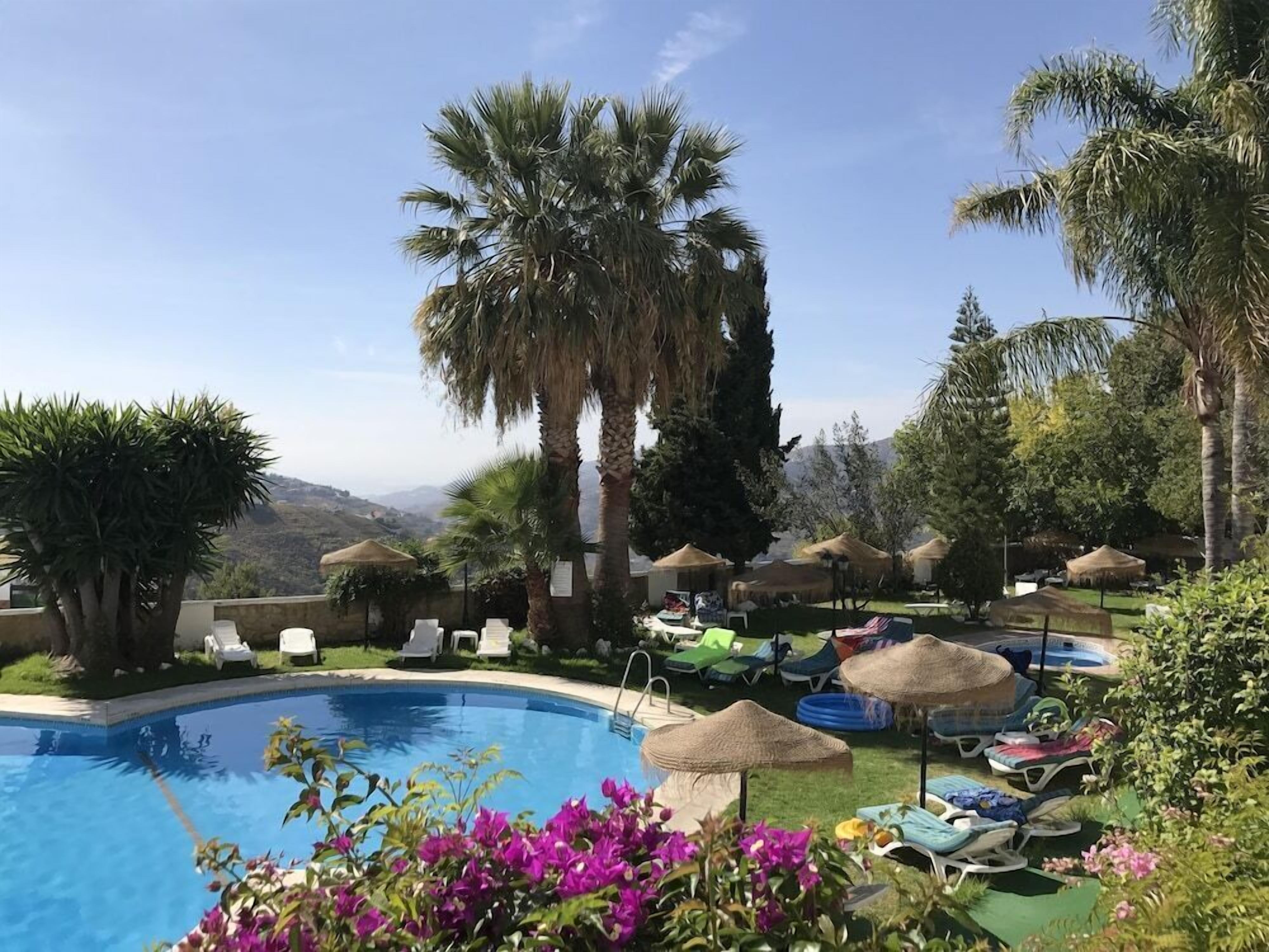 Un espace piscine agrémenté de chaises longues et de parasols, entouré de palmiers et dominé par un paysage montagneux.