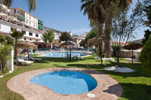 Una piscina con borde de ladrillo, rodeada de césped y palmeras, con edificios al fondo.