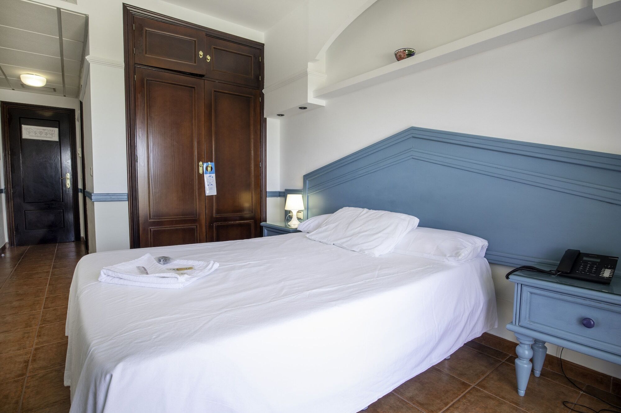 A hotel room with a bed, a nightstand with a phone, a wooden wardrobe, and a door.