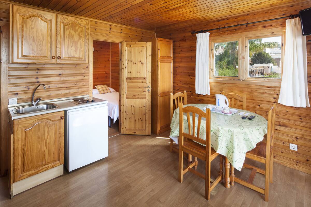Una cabaña de madera con una zona de cocina que incluye un lavabo, un refrigerador pequeño y una mesa de comedor con sillas.