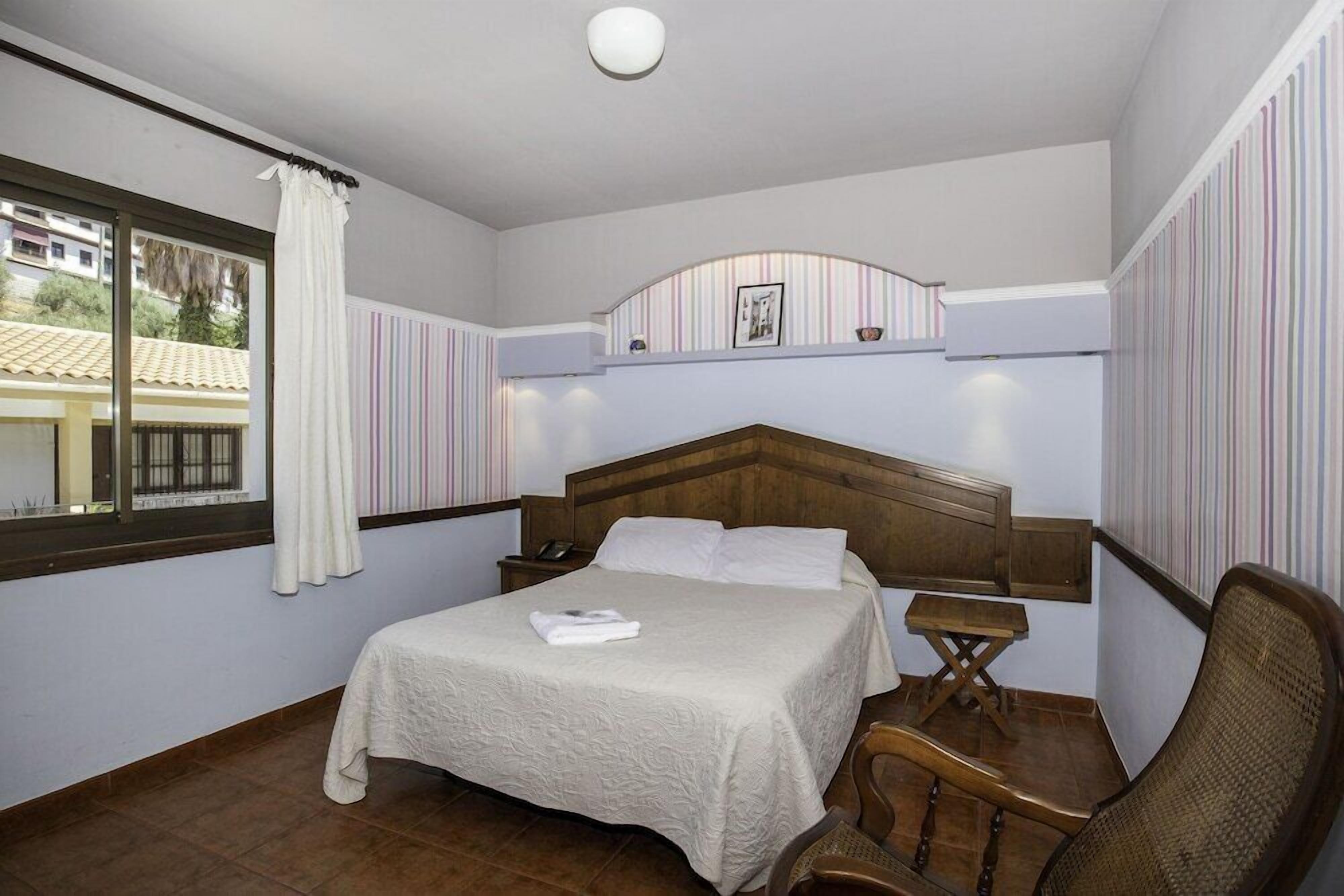 A bedroom with a bed, bedside tables, a chair, a window with curtains, and a view of buildings outside.