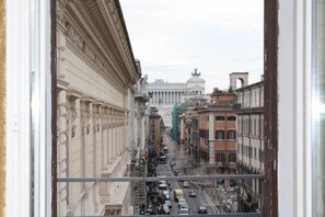 A view from a window overlooking a street with classical architecture, including columns and statues, and a large building with a dome in the background. - Suite Montecitorio (Rome)