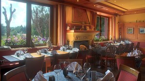 Um restaurante com mesas postas para refeições, uma lareira com a inscrição 'VELLEDA' e amplas janelas com vista para árvores e flores.