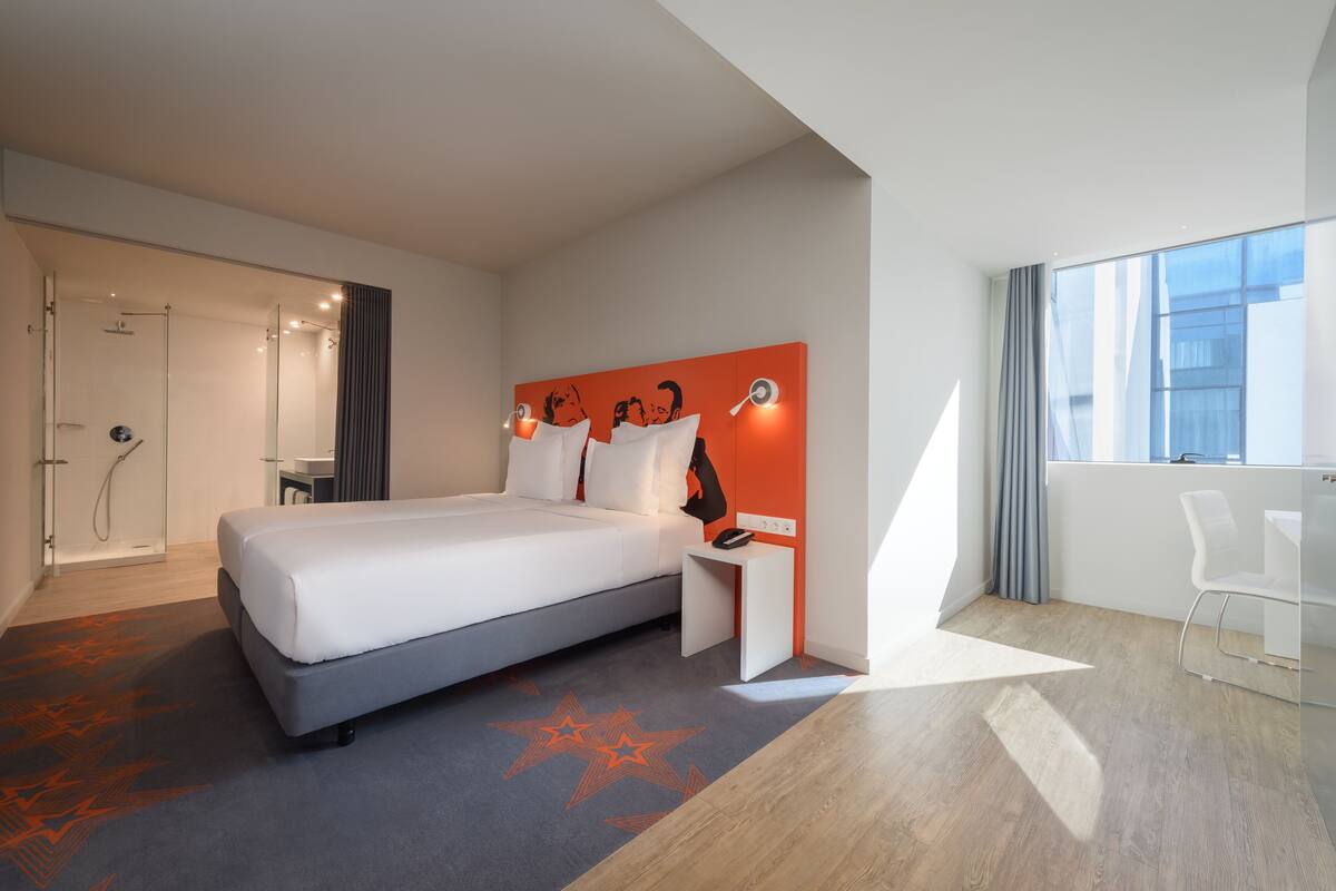 Moderni hotellihuone, jossa on sänky, yöpöytä ja suuri ikkuna.
