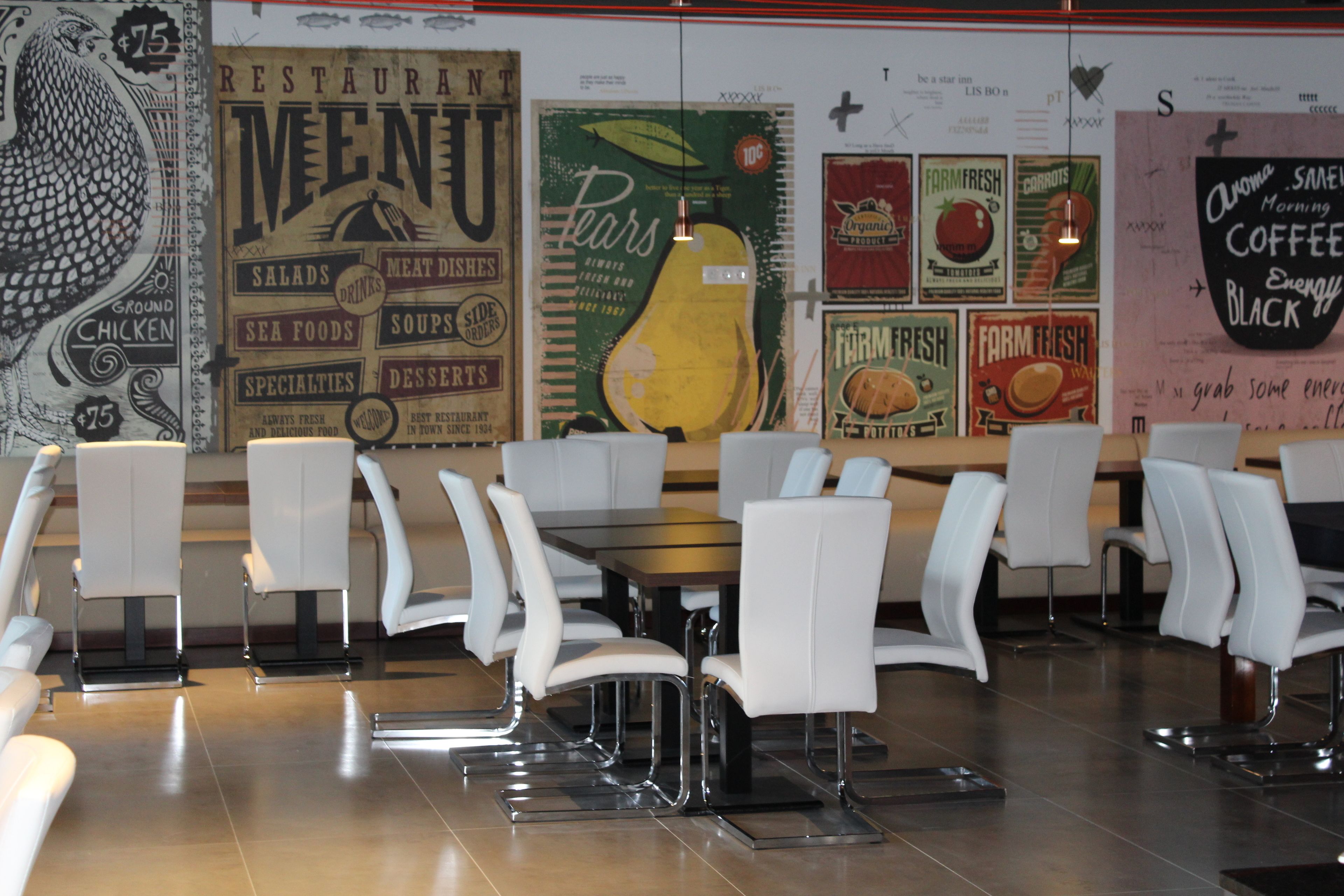 A restaurant interior with white chairs and tables, a menu board, and a wall with various food-related posters.