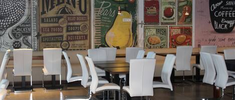 A restaurant interior with white chairs and tables, a menu board, and a wall with various food-related posters.