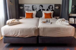 Un letto matrimoniale con lenzuola bianche e cuscini arancioni, un comodino con un vassoio di prodotti per la colazione e un’ampia opera d’arte appesa sopra il letto.