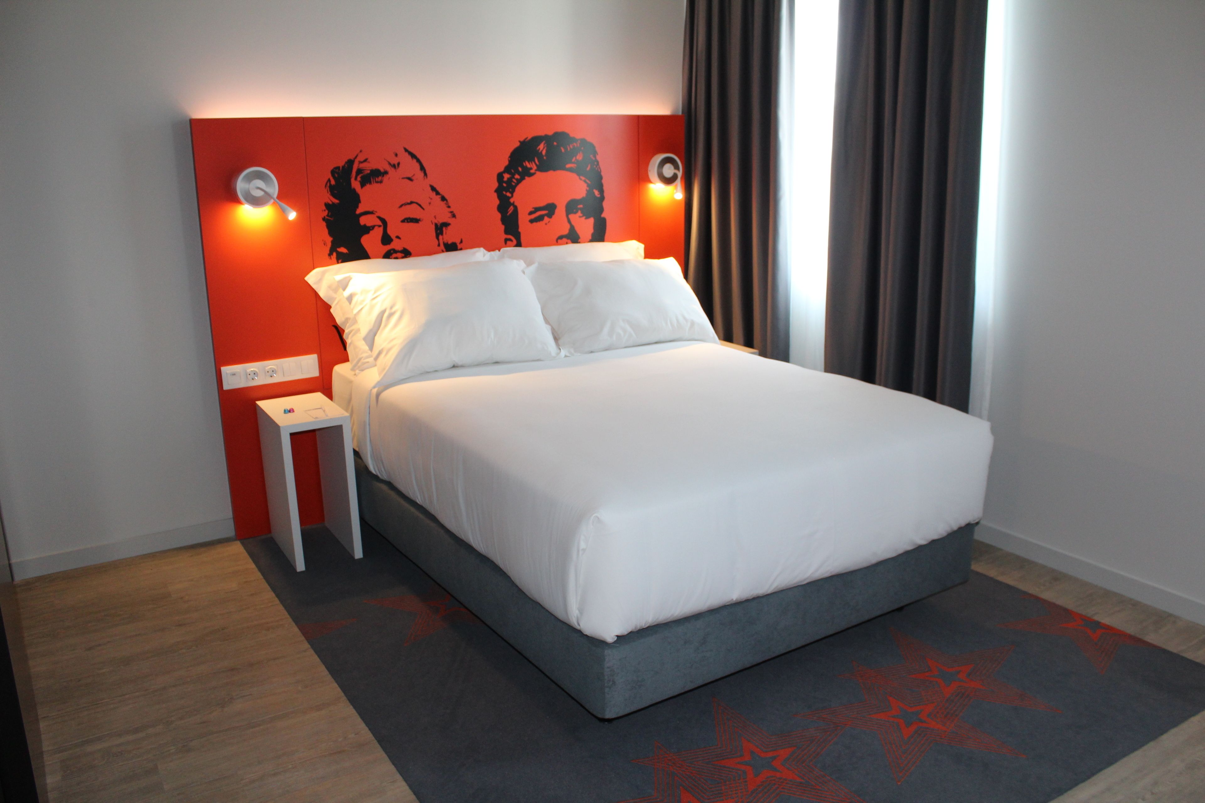 A modern hotel room with a large bed, a small bedside table, and a headboard with a red accent wall.