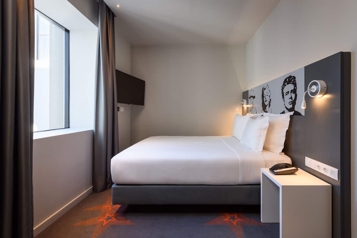Moderni hotellihuone, jossa on suuri sänky, yöpöytä, jossa on puhelin, ja seinälle asennettu televisio.