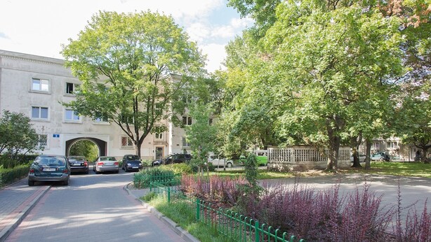 Una calle residencial con coches aparcados, un edificio con un cartel con el número '3' y una valla verde adornada con plantas en flor.