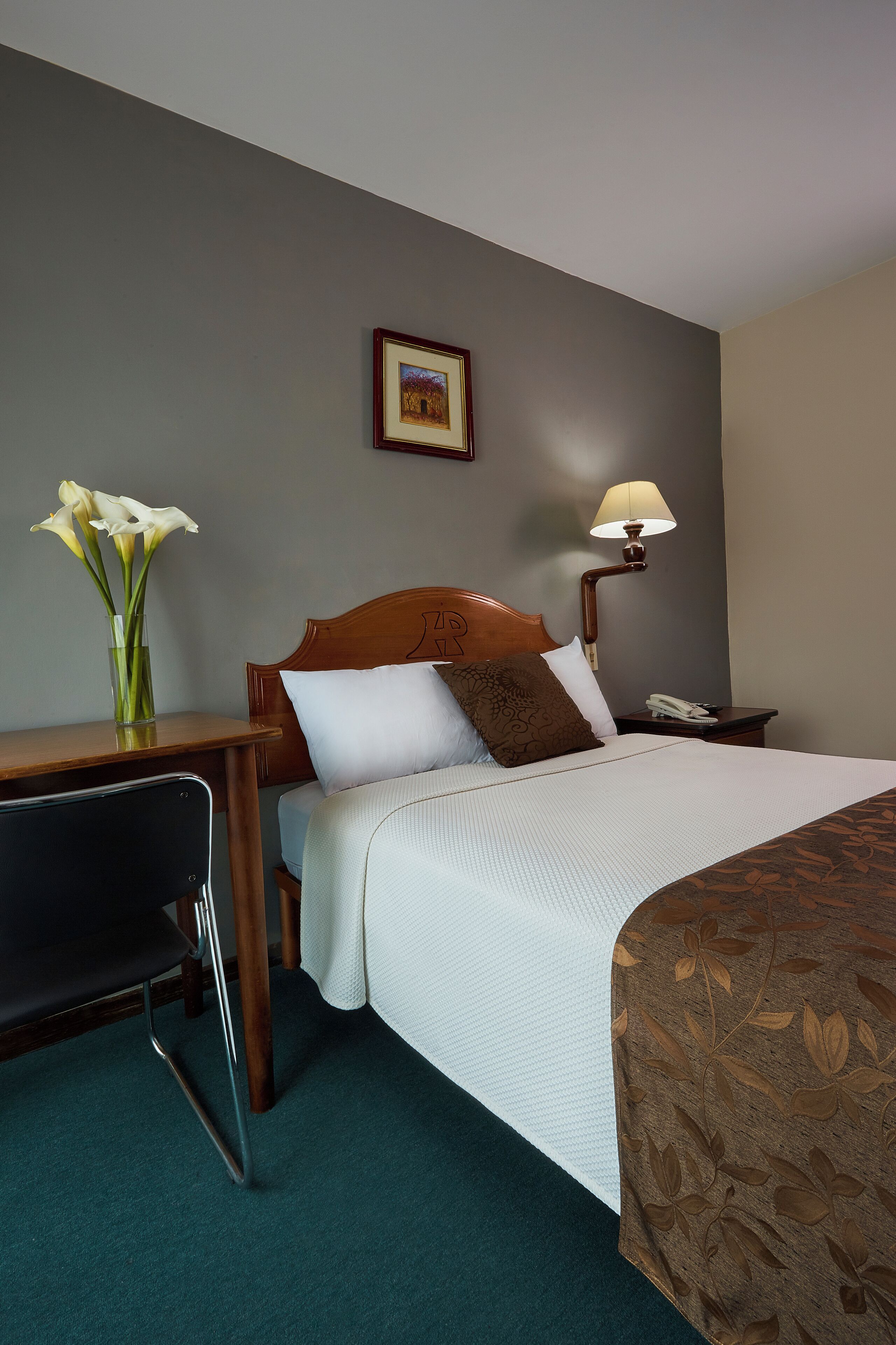 Habitació d'hotel amb llit, tauleta de nit, làmpada, cadira i un vas amb flors.