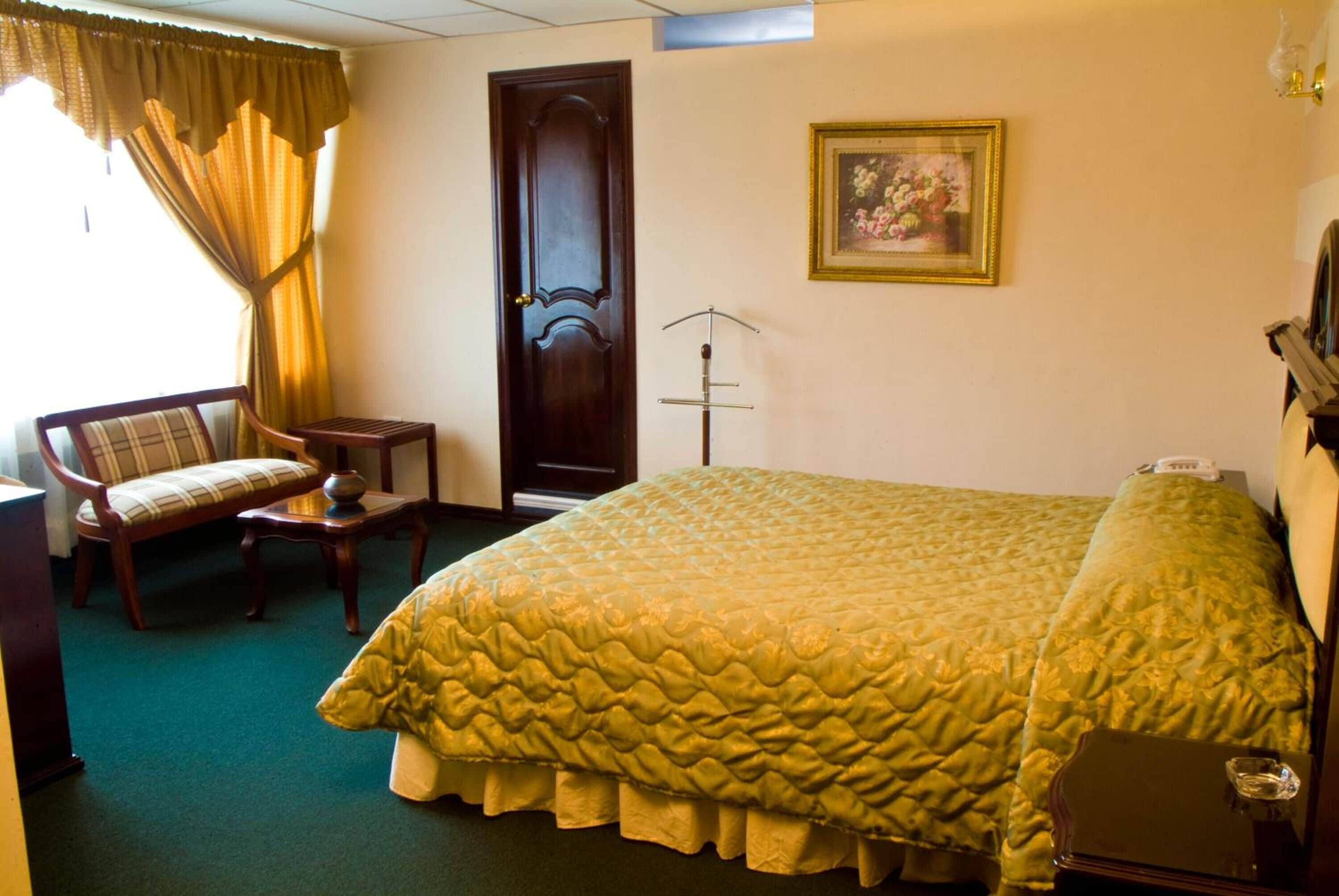 A hotel room with a large bed, a chair, a small table, a door, and a framed picture on the wall.