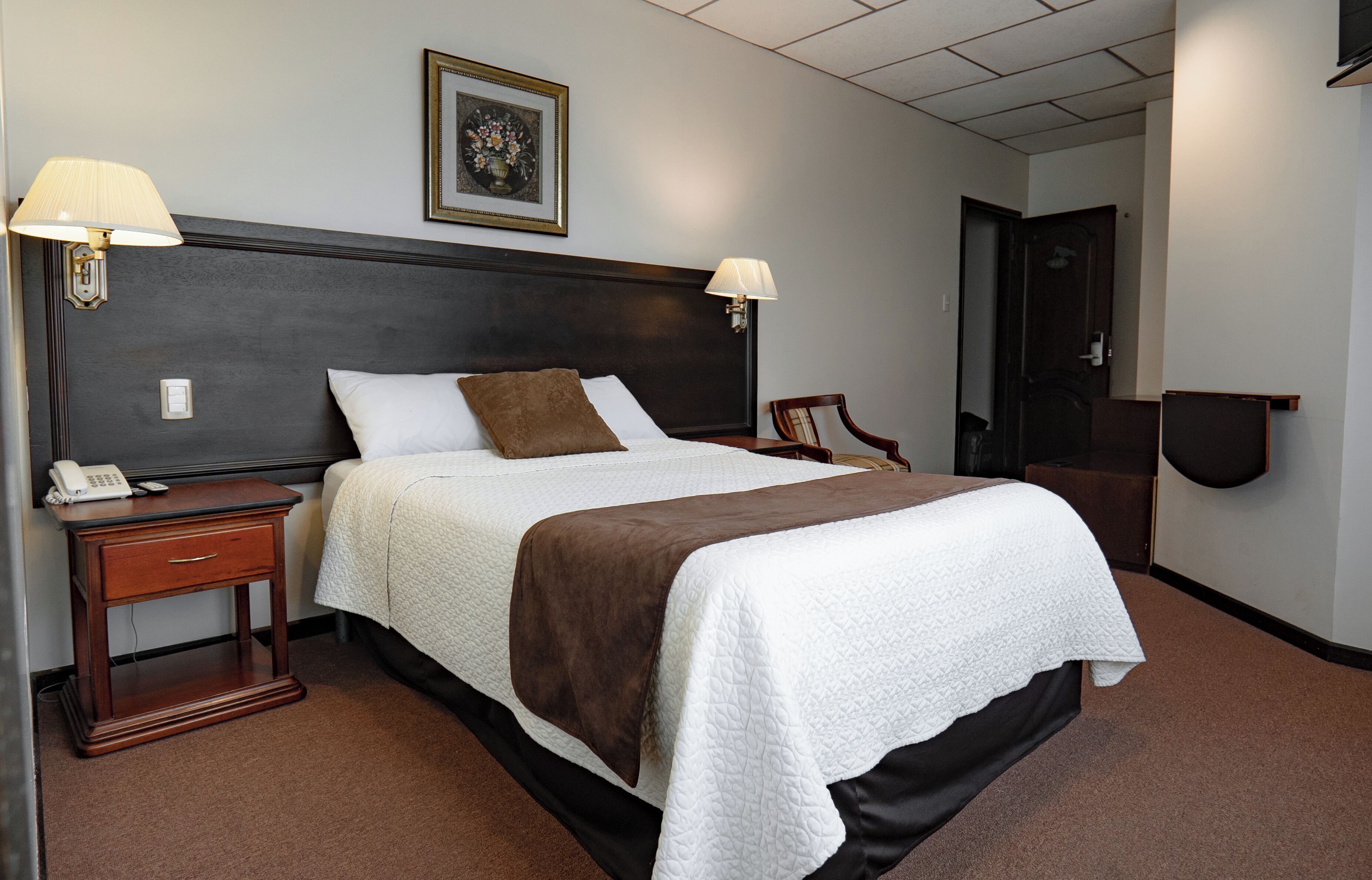 Una habitación de hotel con una cama grande, dos lámparas de noche, una mesita de noche con un teléfono, un cuadro en la pared y una puerta a la derecha.