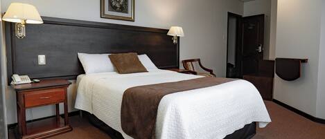 Una habitación de hotel con una cama grande, dos lámparas de noche, una mesita de noche con un teléfono, un cuadro en la pared y una puerta a la derecha.