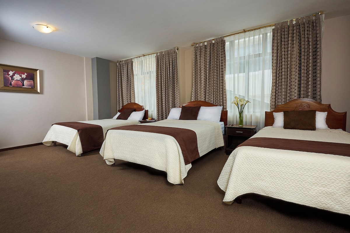 A hotel room with three beds, each with a brown headboard and white bedding. There is a framed picture on the wall, a nightstand with a vase of flowers, and a window with patterned curtains.