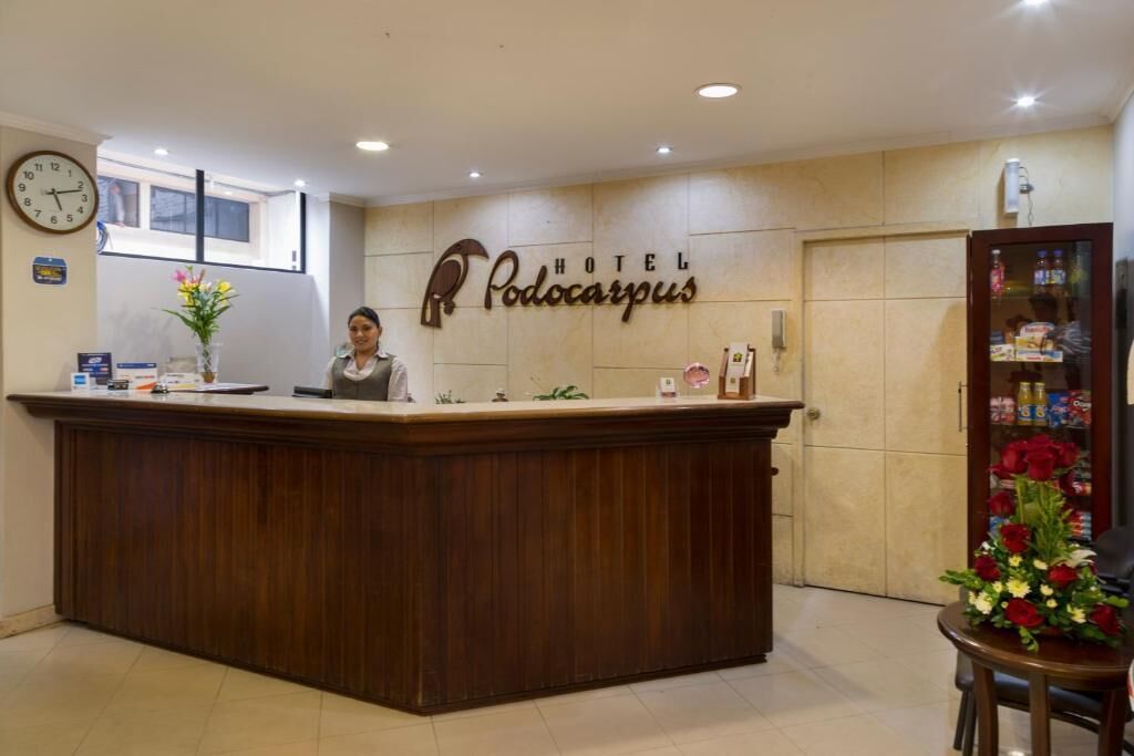 A reception desk with a clock, a sign reading "HOTEL Podocarpus," and a flower arrangement.