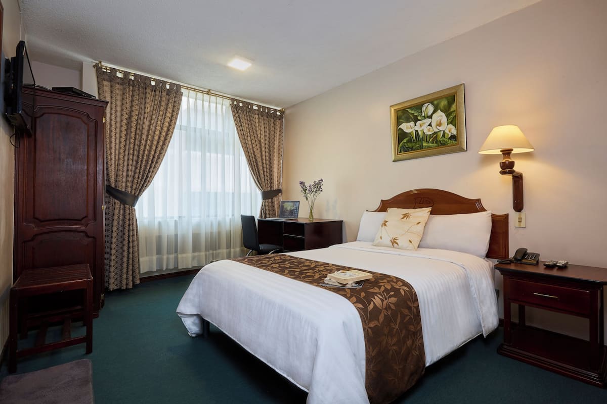 A hotel room with a large bed, a desk, a chair, a wardrobe, a painting on the wall, and a lamp.