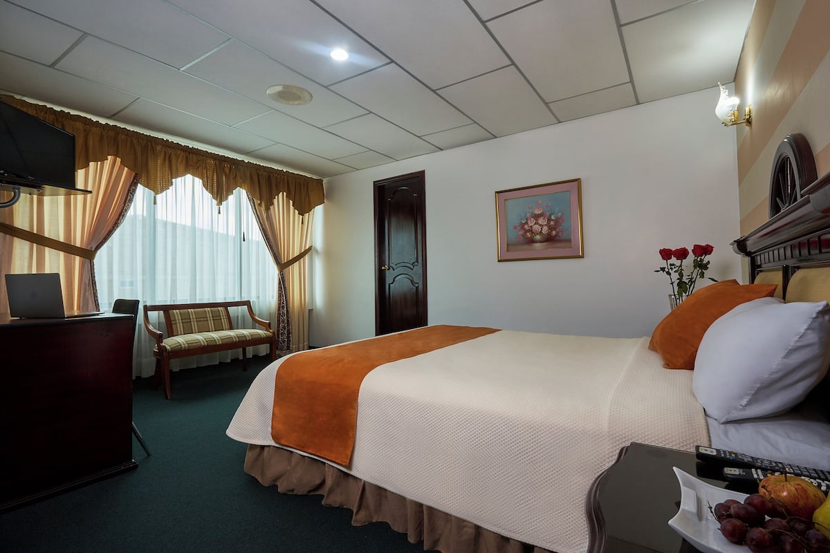 A hotel room with a bed, a desk, a chair, a TV, and a painting on the wall.