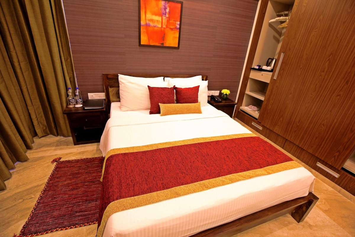Une chambre d’hôtel avec un grand lit, une armoire en bois, une table de chevet avec un téléphone et un vase de fleurs, ainsi qu’un tapis rouge et or à rayures.