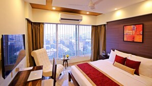 Habitación de hotel con una cama grande, un escritorio con una computadora portátil, una silla y vistas de la ciudad a través de grandes ventanales.