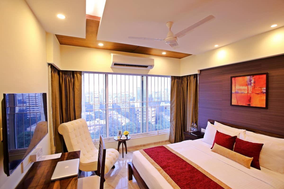 Une chambre d’hôtel avec un grand lit, un bureau avec un ordinateur portable, une chaise et une vue sur la ville grâce à de grandes fenêtres.
