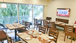 Un restaurante con varias mesas y sillas, amplias ventanas que dan a zonas verdes y un televisor montado en la pared.