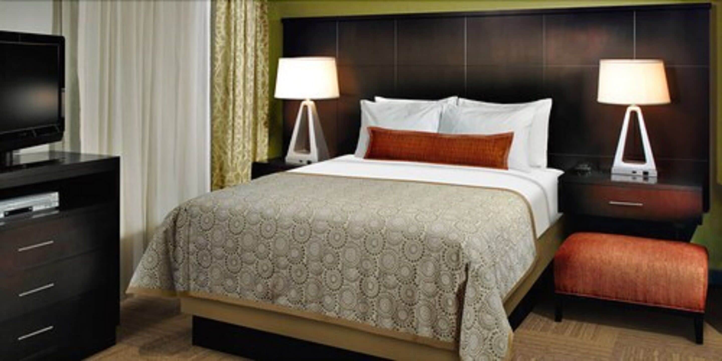 Ein Hotelzimmer mit einem Bett, zwei Nachttischen mit Lampen, einem Fernseher, einem Schminktisch und einem Hocker.
