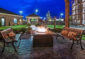 Un patio al aire libre con piso de piedra, dos bancos de madera, una chimenea central y un pabellón al fondo.