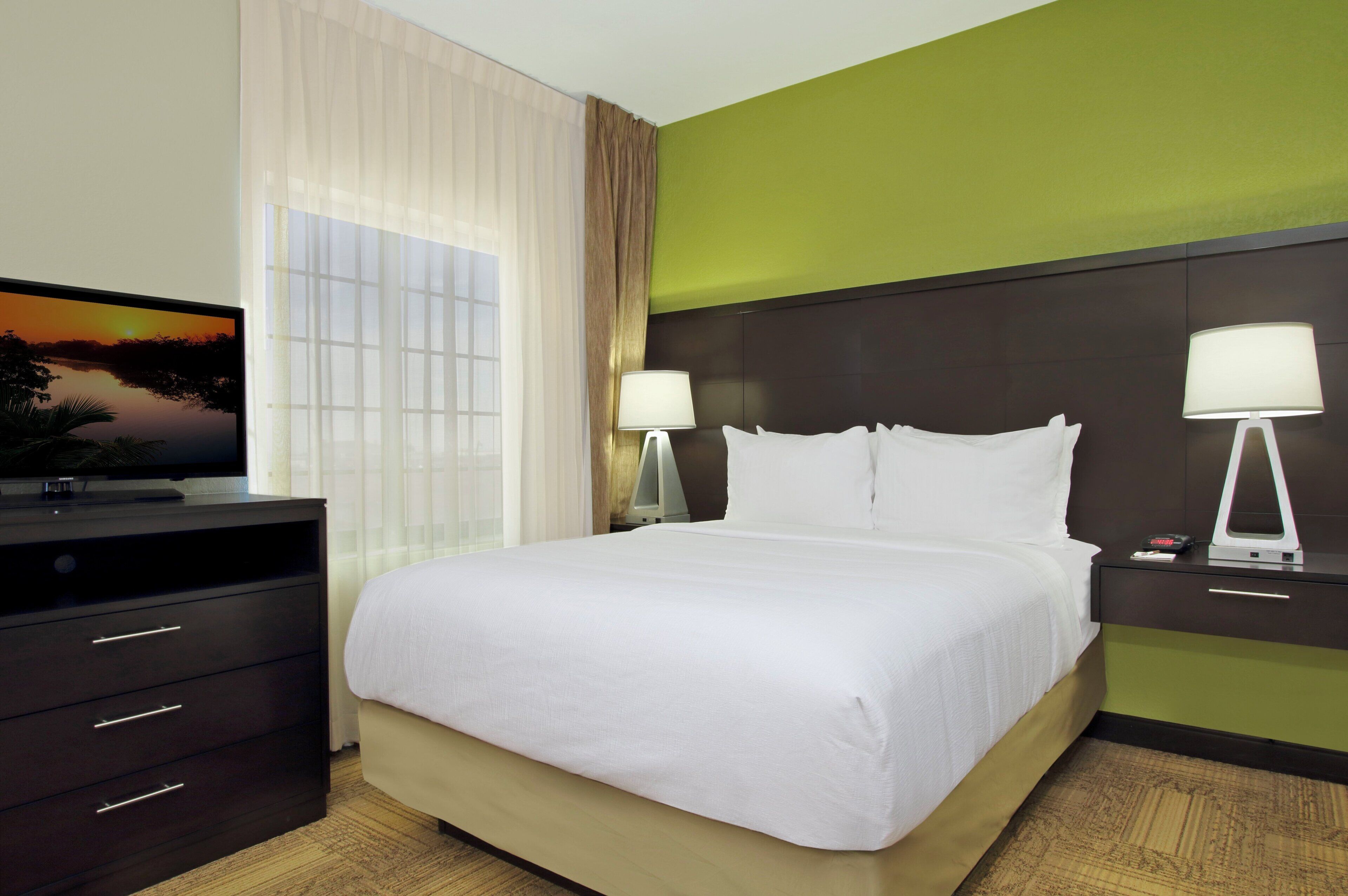 A hotel room with a bed, two bedside lamps, a TV on a dresser, and a painting of a sunset.